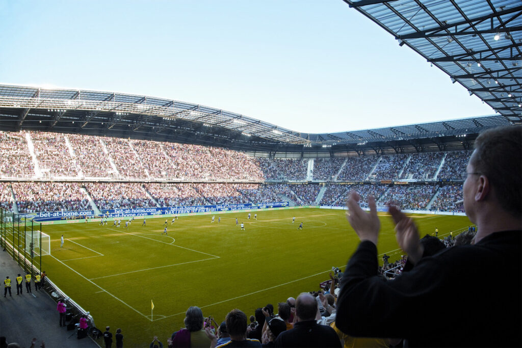 Volles Stadion bei Sonnenschein. Im Vodergurnd sieht man einen Mann klatschen und auf das Spielfeld schauen.